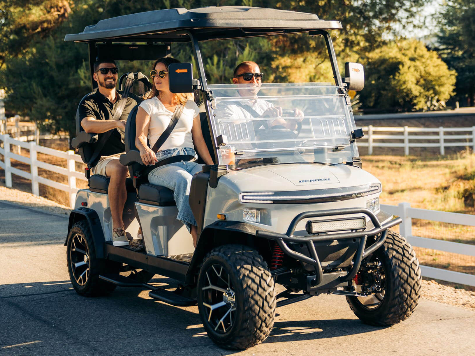 03 Best Golf Cart 2026 Denago Rover XL Three friends riding a lifted white 2026 Denago Rover XL golf cart down a country road at sunset